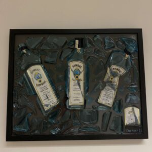 Een zwart omlijst kunstwerk met drie Bombay Sapphire gin flessen ingebed in een gebroken glazen oppervlak.